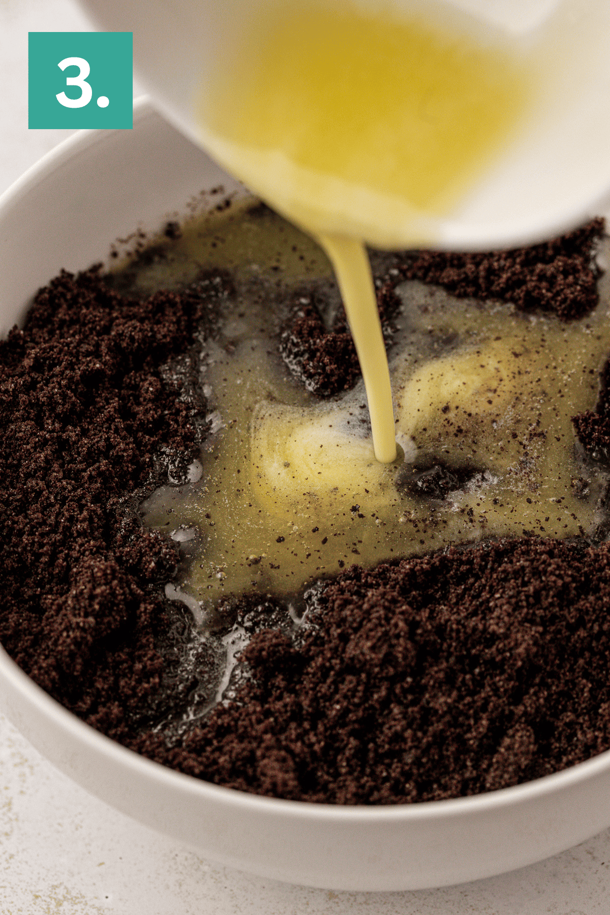 Step 3: Melted butter being poured into a bowl of crushed chocolate cookies to form an Oreo pie crust, perfect as the base for your next delicious dessert.