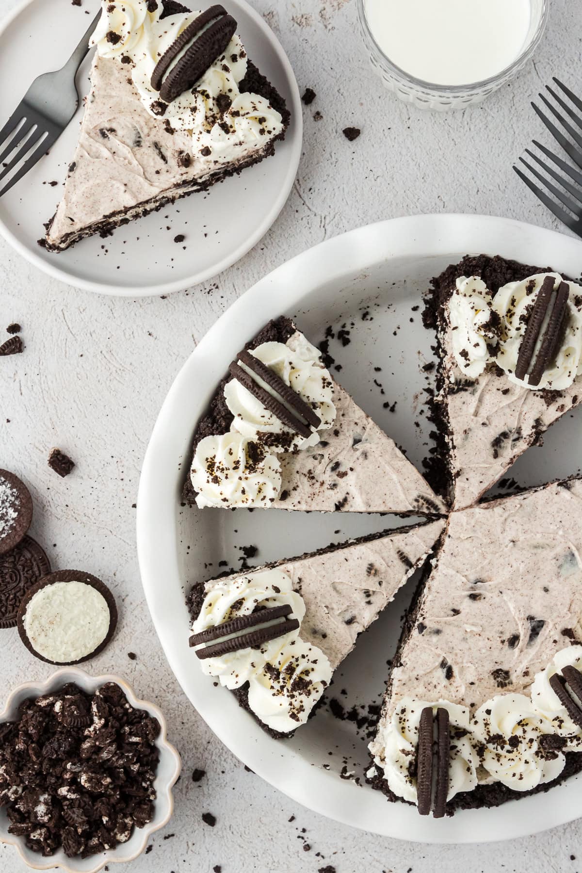 Top-down view of a cookies and cream pie with an Oreo pie crust, sliced and topped with whipped cream and mini cookies, a slice served on a plate, and a glass of milk nearby on a light surface.