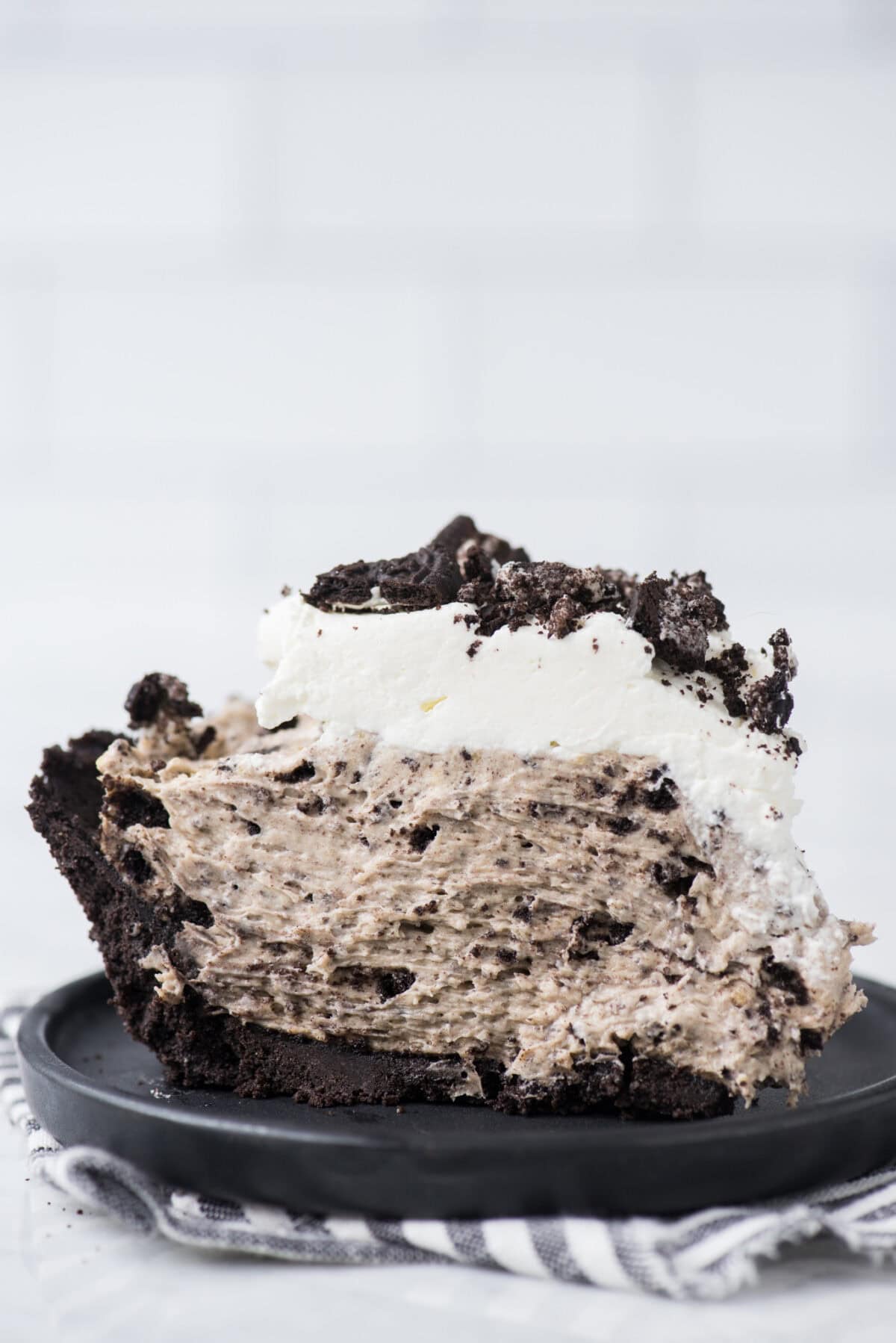 A close-up of a slice of cookies and cream pie with a crumbly Oreo pie crust, creamy filling mixed with cookie pieces, and topped with whipped cream and more cookie crumbles, served on a black plate.