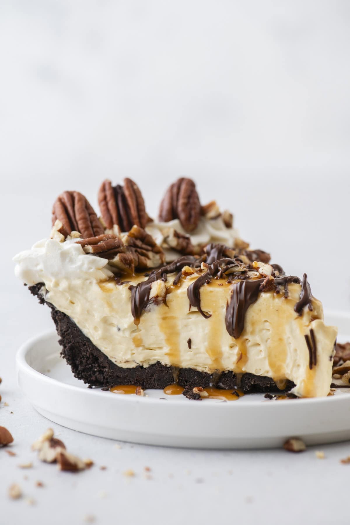 A slice of creamy pie with an Oreo pie crust, topped with whipped cream, pecans, caramel, and chocolate drizzle, sits on a white plate with scattered pecans around it.