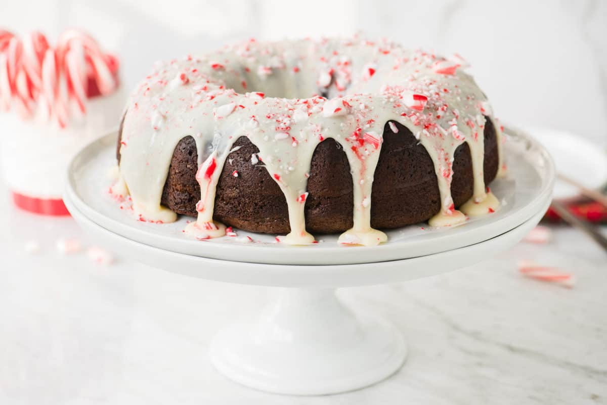 whole chocolate bundt cake with white chocolate glaze and candy cane pieces on white cake stand