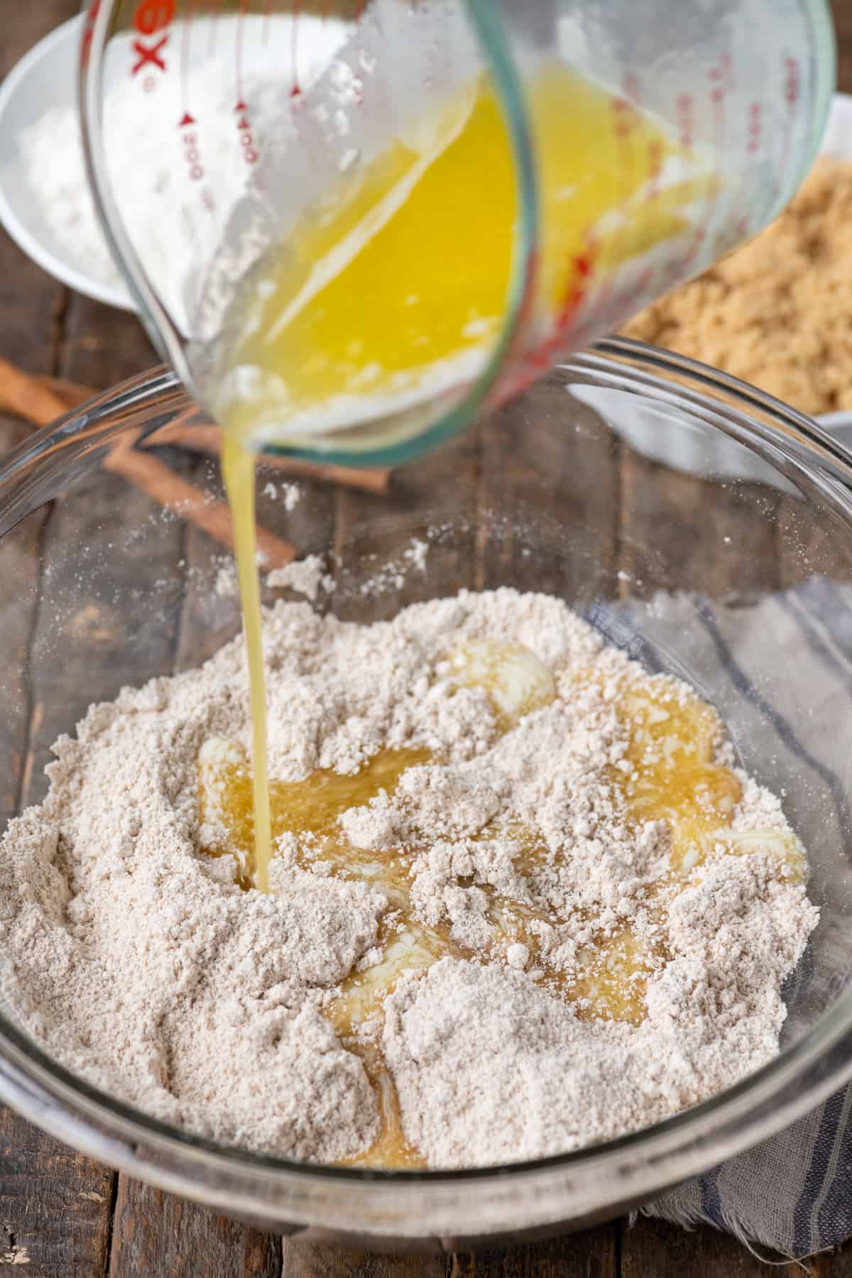 melted butter being poured into glass bowl with crumb topping mixture on wood background