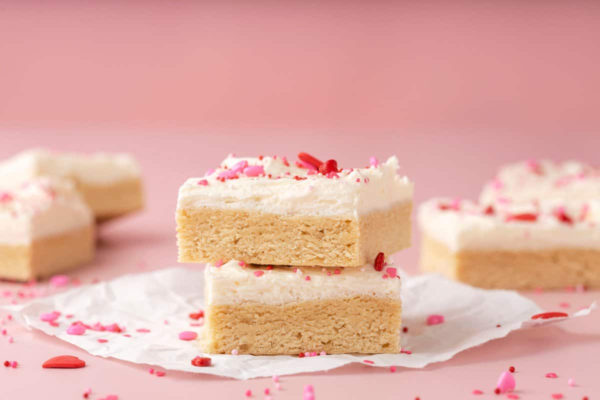 two sugar cookie bars with white frosting and valentine’s sprinkles on pink background