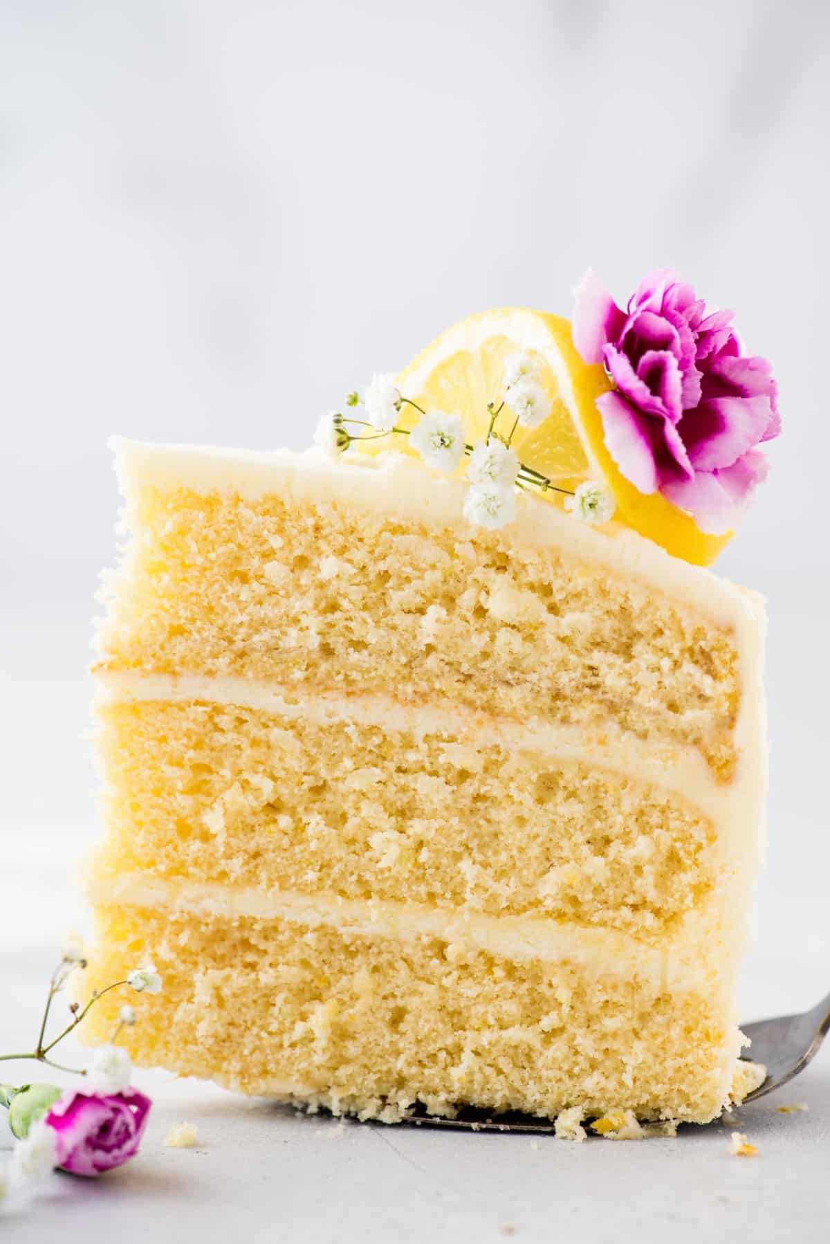 Slice of lemon layer cake on cake server, garnished with flowers and fresh lemon