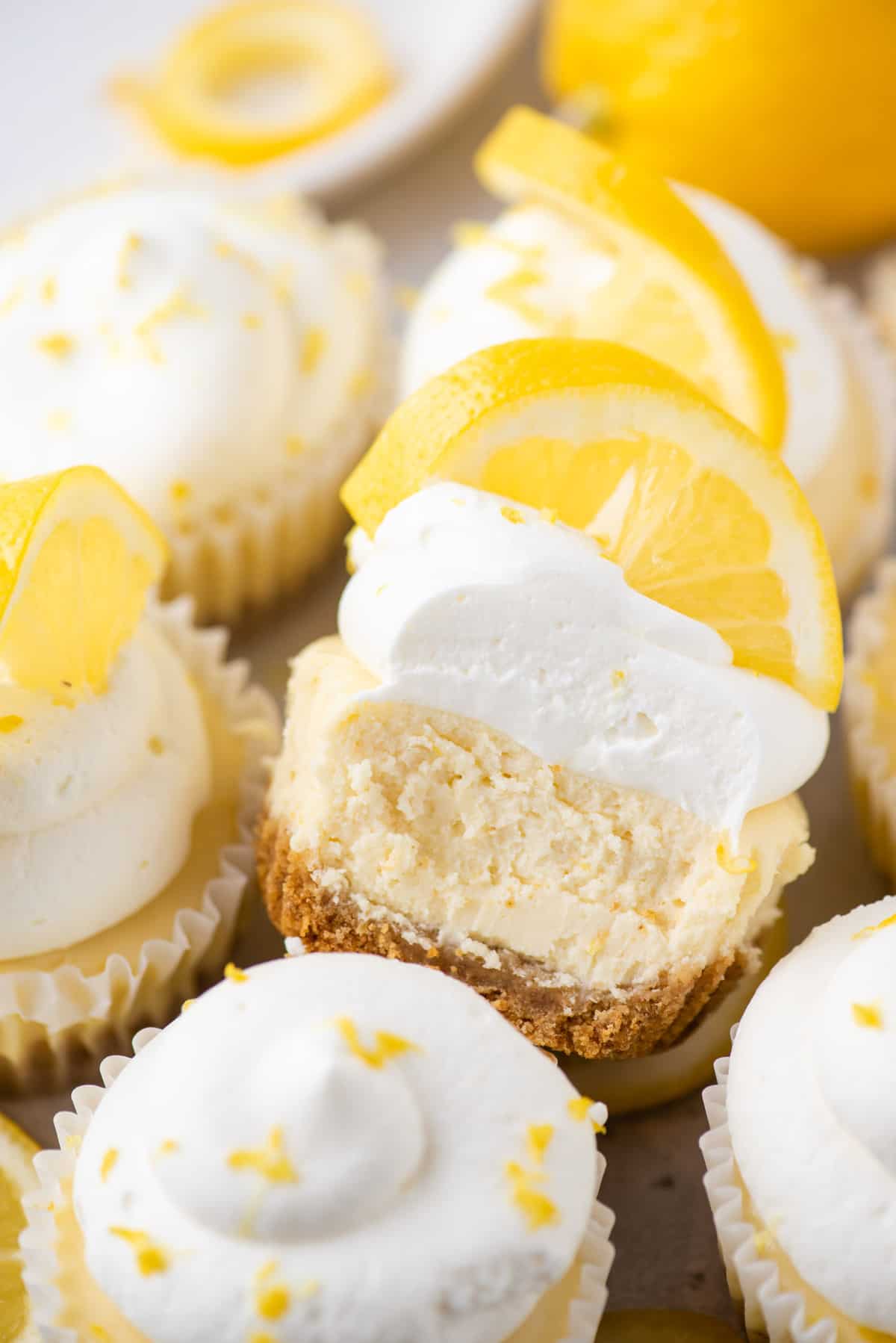 Mini lemon cheesecake topped with whipped cream and lemon slice with bite removed with more mini cheesecakes surrounding it