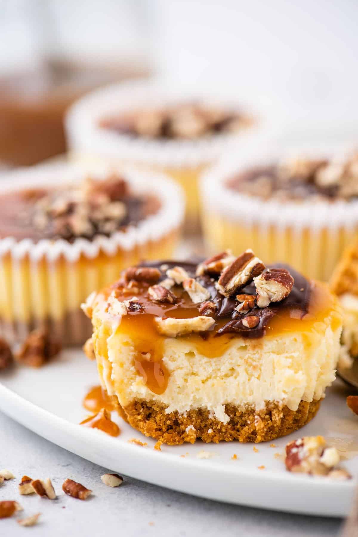 mini cheesecake topped with caramel, chocolate and chopped pecans with bite removed on white plate