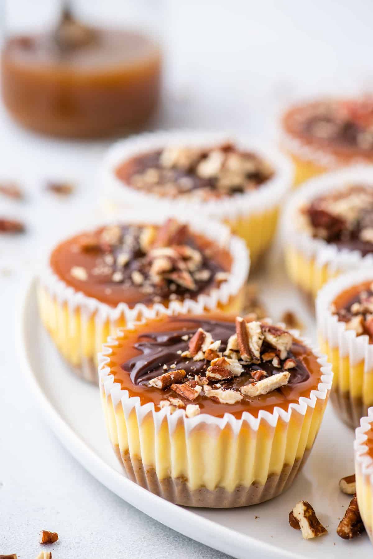 mini cheesecakes topped with caramel, chocolate and chopped pecans arranged on white tray