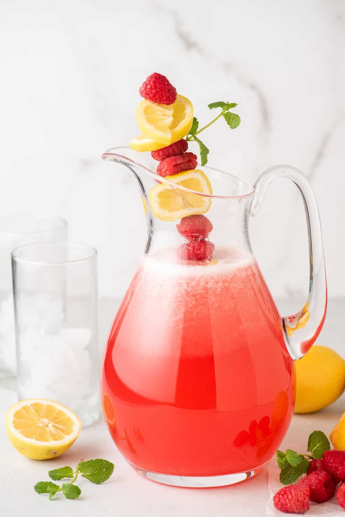Pitcher of raspberry lemonade garnished with skewer of raspberries, lemon slices, and sprig of mint