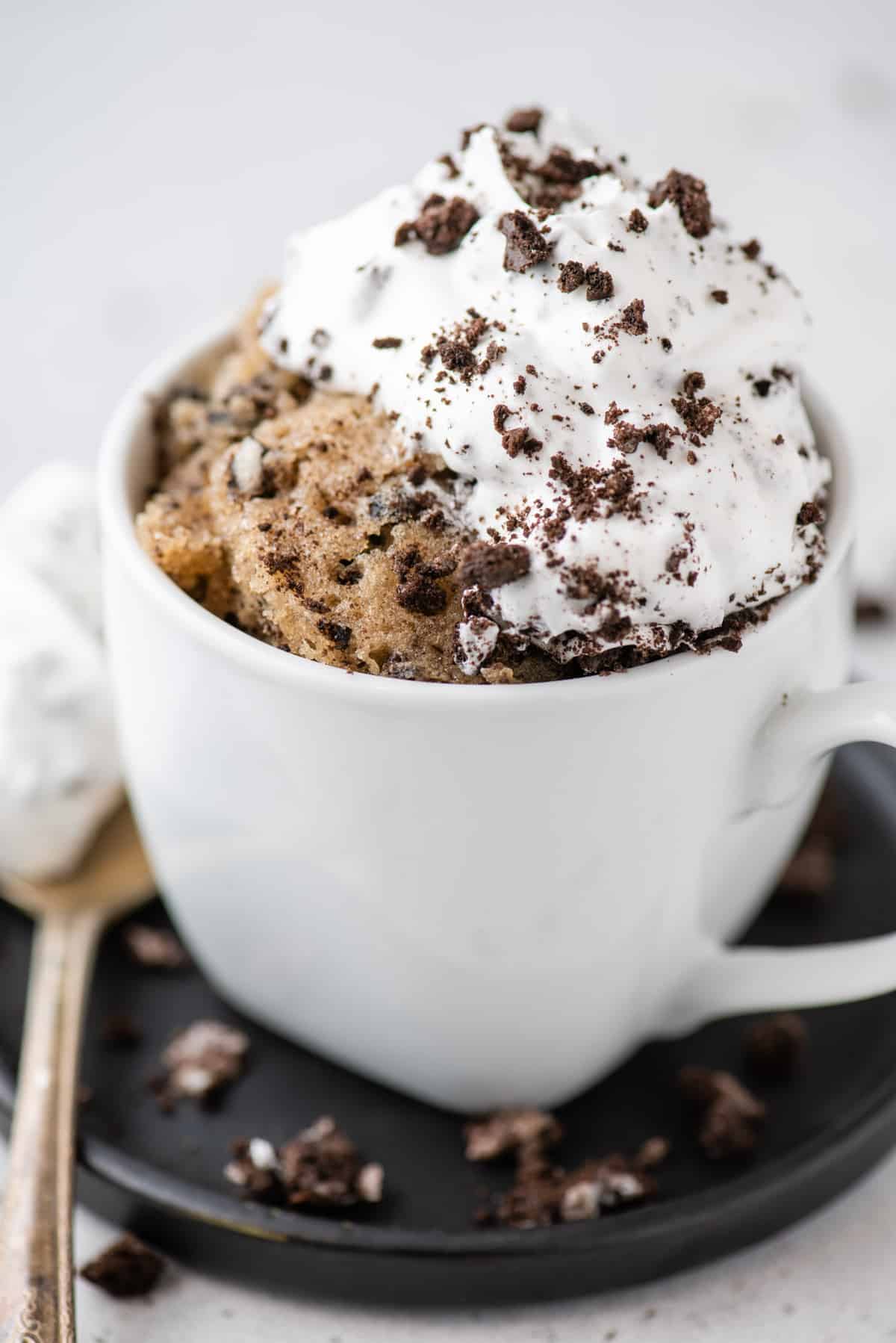 Oreo mug cake in white mug set on black plate, topped with whipped cream and crushed Oreos