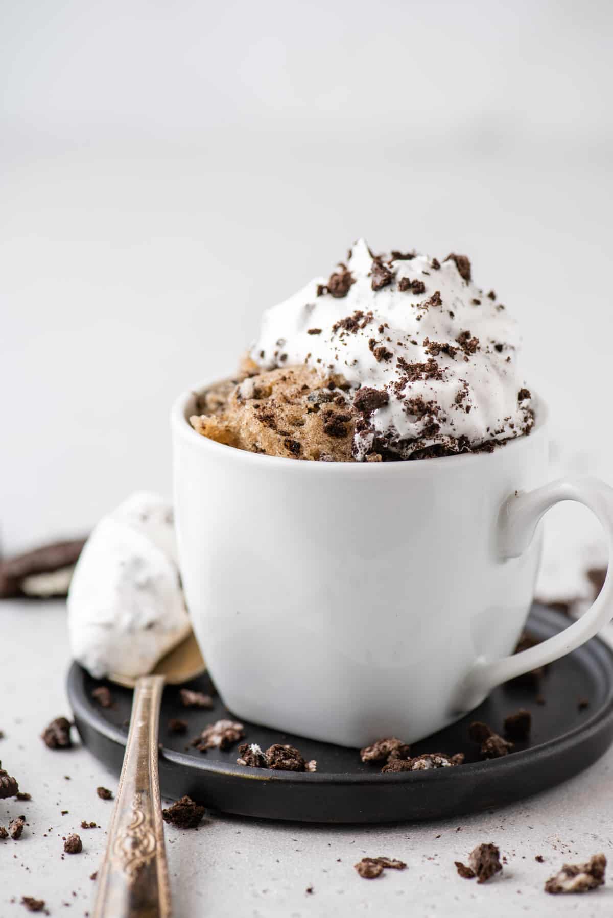 Oreo mug cake in white mug, set on black plate with spoon of whipped cream