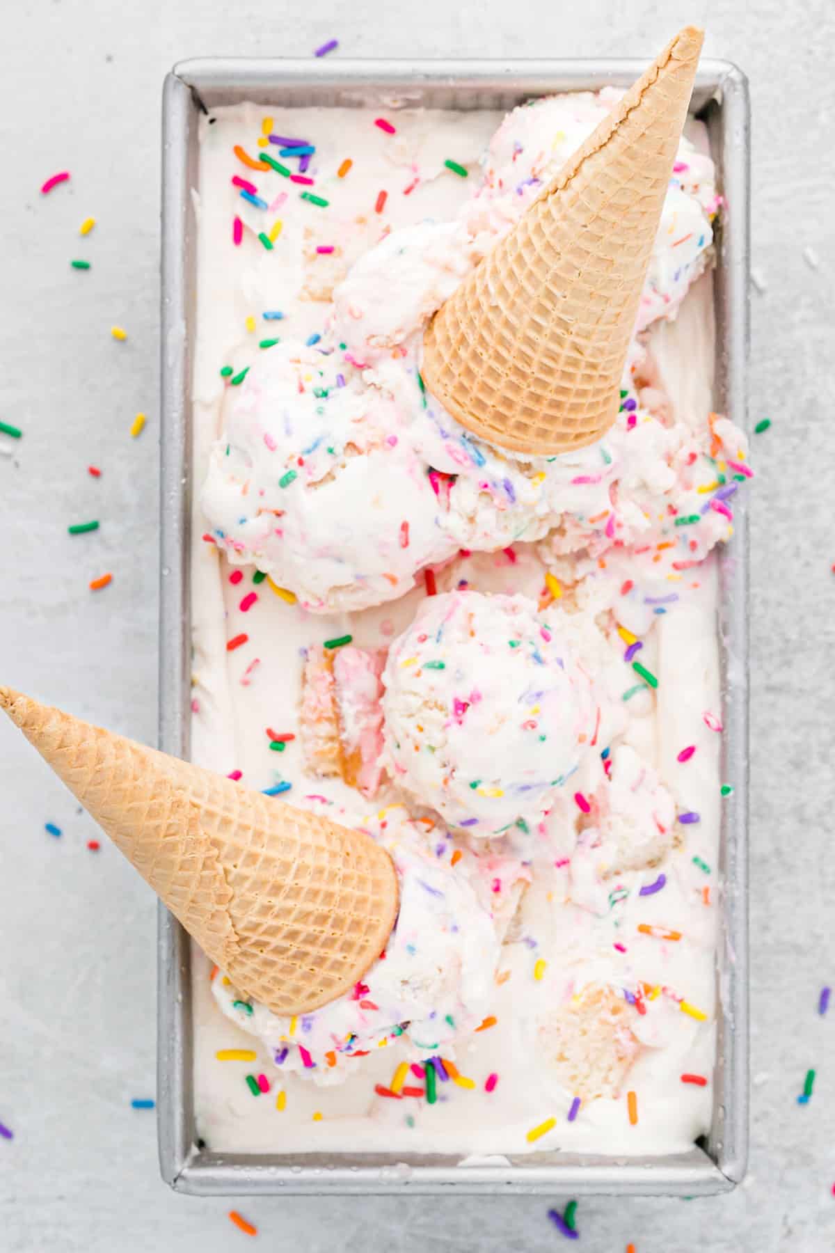 Loaf pan of birthday cake ice cream with scoops and cones on top