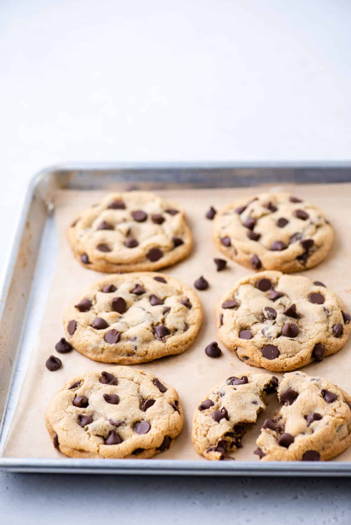 6 chocolate chip cookies on a parchment-lined baking sheet, with one broken in half