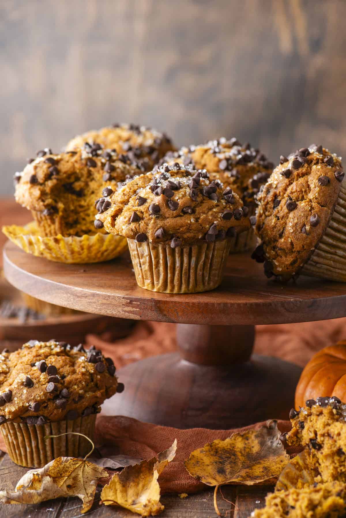 Pumpkin Chocolate Chip Muffins sit on a wooden cake stand, topped with mini chocolate chips. Autumn leaves and a small pumpkin surround the display, perfectly capturing the cozy fall atmosphere.