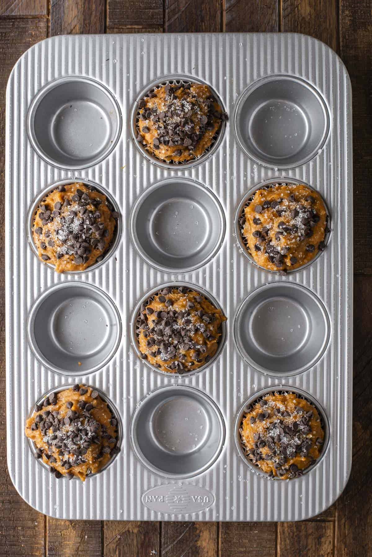 A muffin pan with eight filled cups of Pumpkin Chocolate Chip Muffins batter, each topped with chocolate chips and sugar, and four empty cups, sits on a wooden surface.