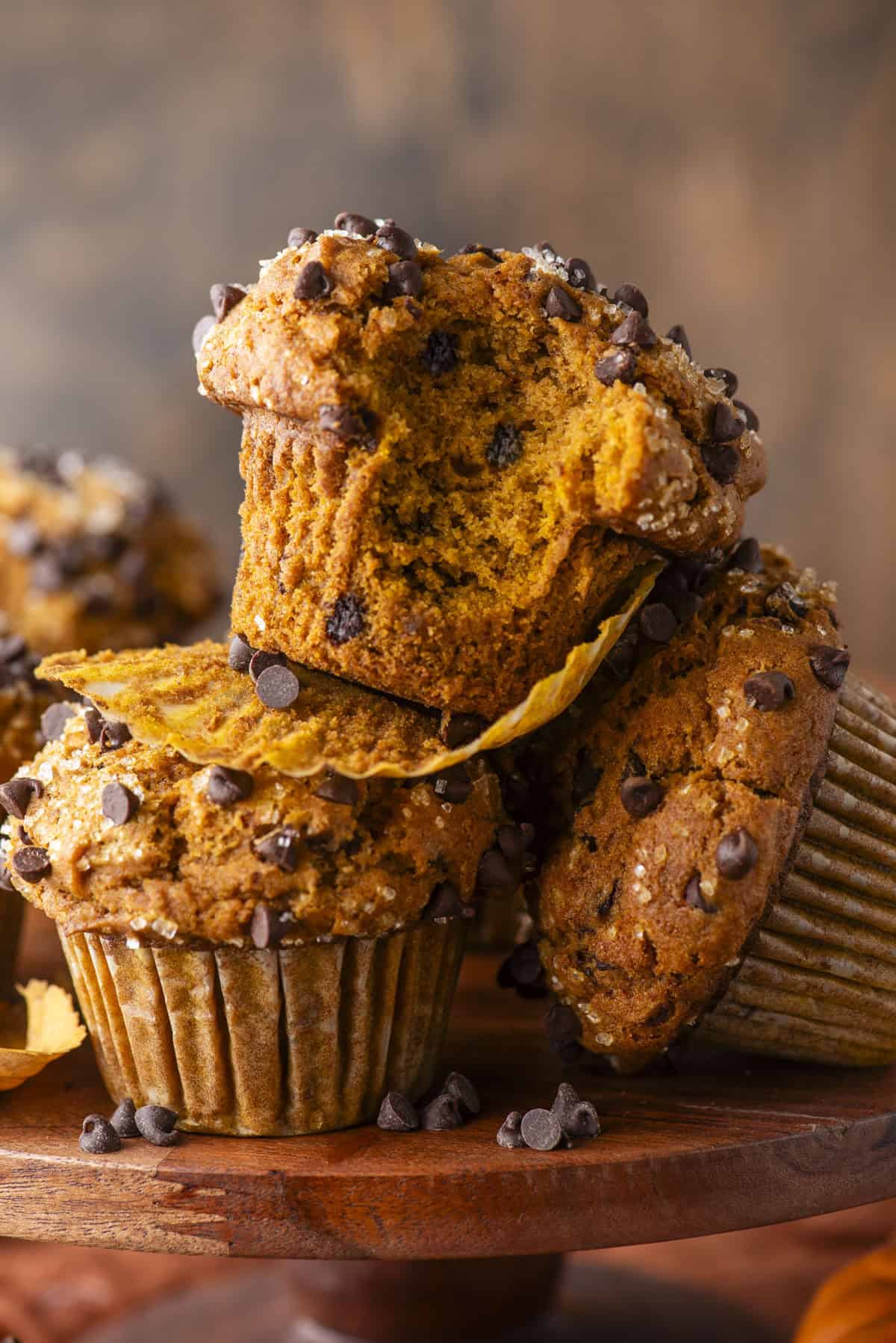 A close-up of Pumpkin Chocolate Chip Muffins, one with a bite taken out, stacked on a wooden stand against a rustic brown background.