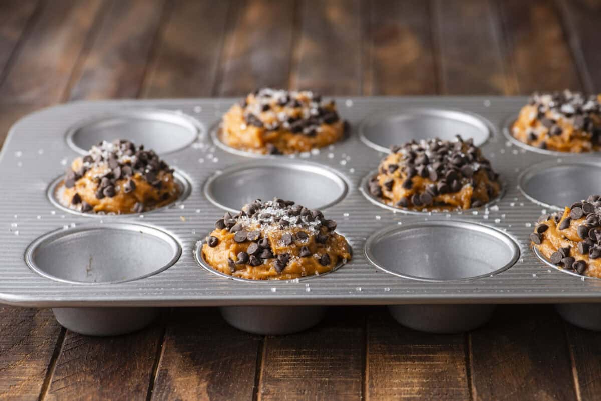 A metal muffin tin holds six unbaked Pumpkin Chocolate Chip Muffins batter mounds topped with chocolate chips and sprinkled with coarse salt, on a rustic wooden surface. The other spaces in the tin are empty.