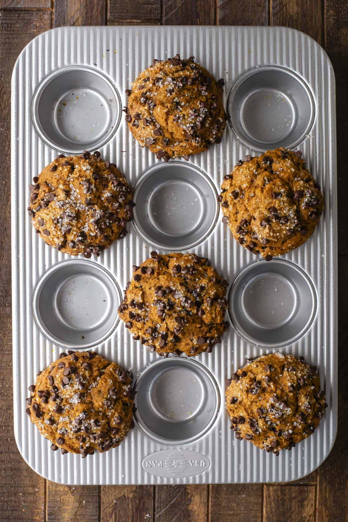 A muffin tin with seven Pumpkin Chocolate Chip Muffins sprinkled with sugar sits on a wooden surface, while five spaces in the tin are empty.