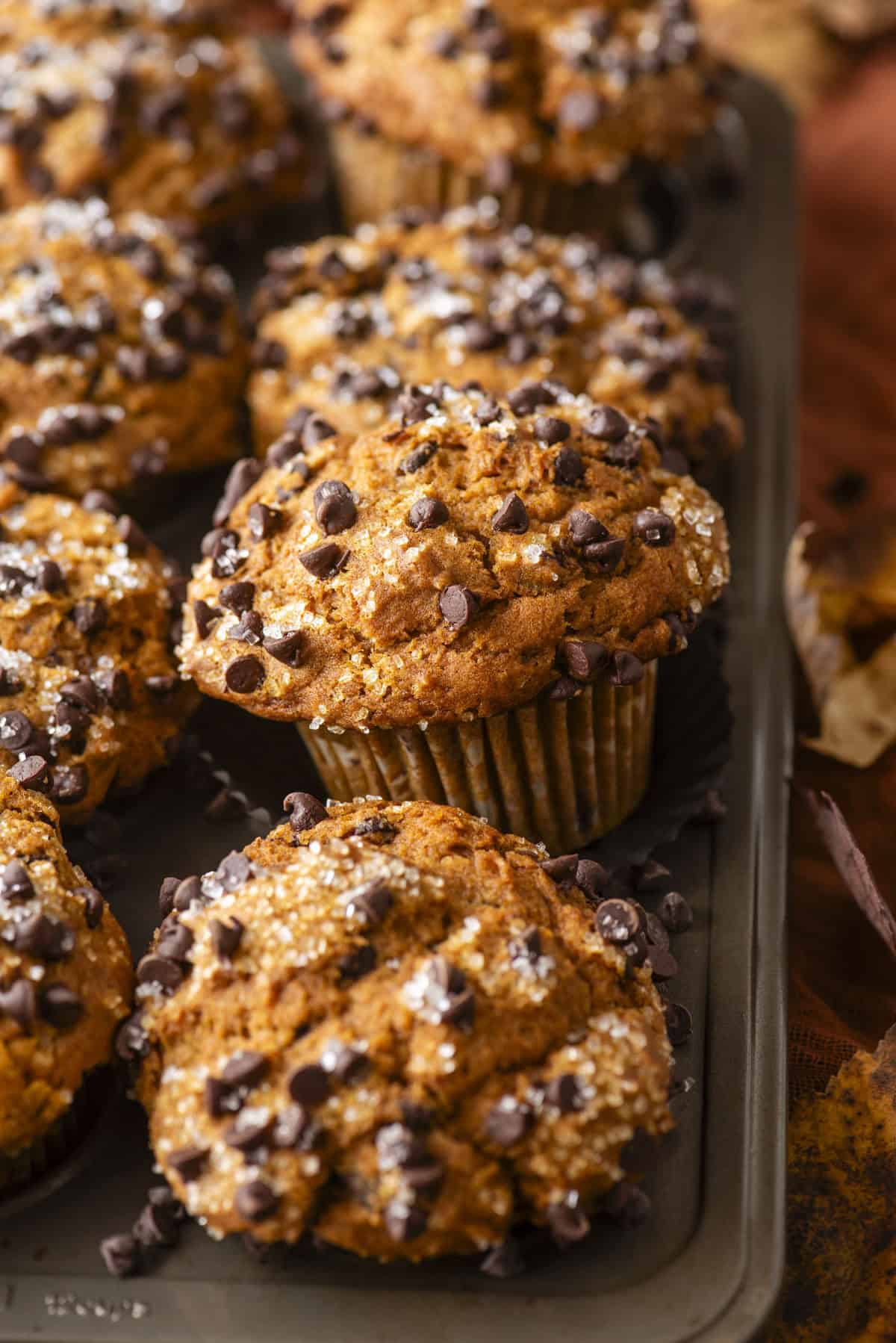 A close-up of a muffin tin filled with Pumpkin Chocolate Chip Muffins, freshly baked and topped with coarse sugar and mini chocolate chips. The muffins have a golden-brown, textured surface.