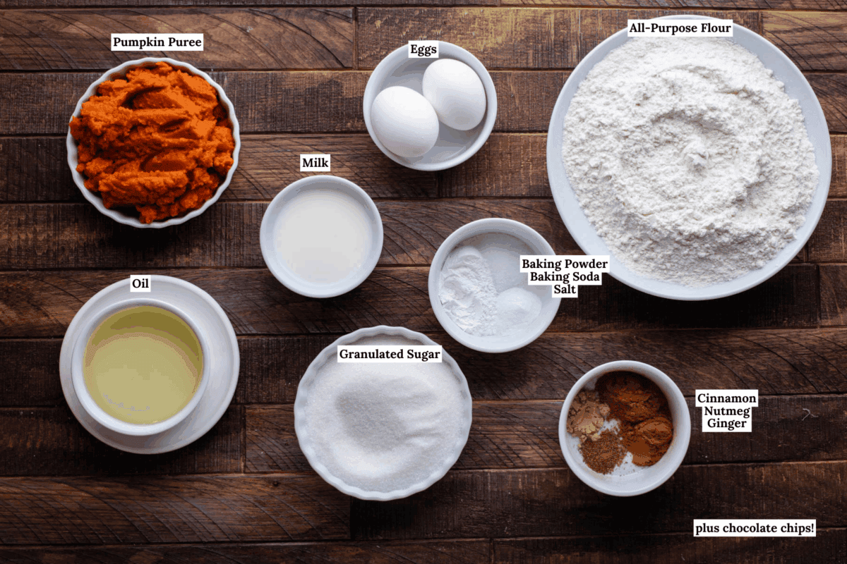 A top-down view of ingredients for Pumpkin Chocolate Chip Muffins on a wooden table: pumpkin puree, eggs, flour, milk, oil, sugar, baking powder, baking soda, salt, cinnamon, nutmeg, ginger, and optional chocolate chips.