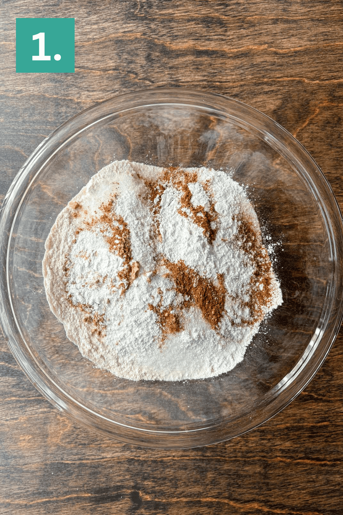 A glass bowl containing flour and spices, key ingredients for Pumpkin Chocolate Chip Muffins, sits on a wooden surface. The image is labeled with a green box and the number 1 in the top left corner.