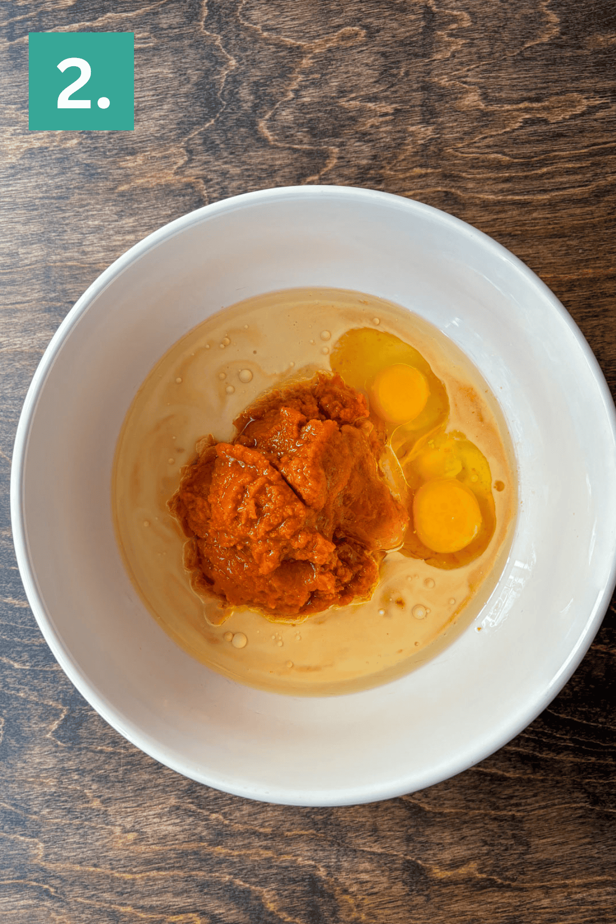 A white bowl on a wooden surface contains two raw eggs and a scoop of orange pumpkin puree, ready to be mixed for Pumpkin Chocolate Chip Muffins. A small green box labeled 2 sits in the top left corner.