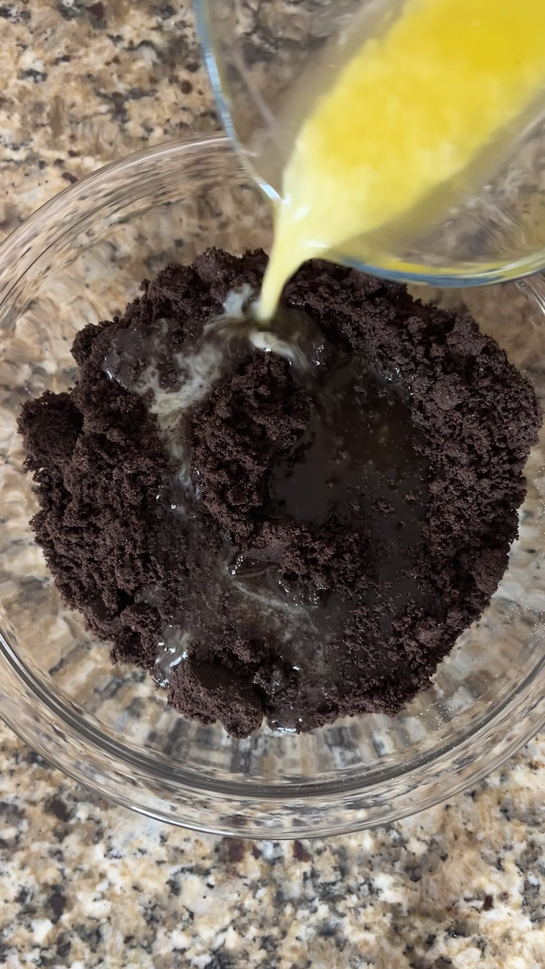 A glass bowl with crushed chocolate cookies for a no bake peanut butter pie has melted butter being poured over them, on a granite countertop.