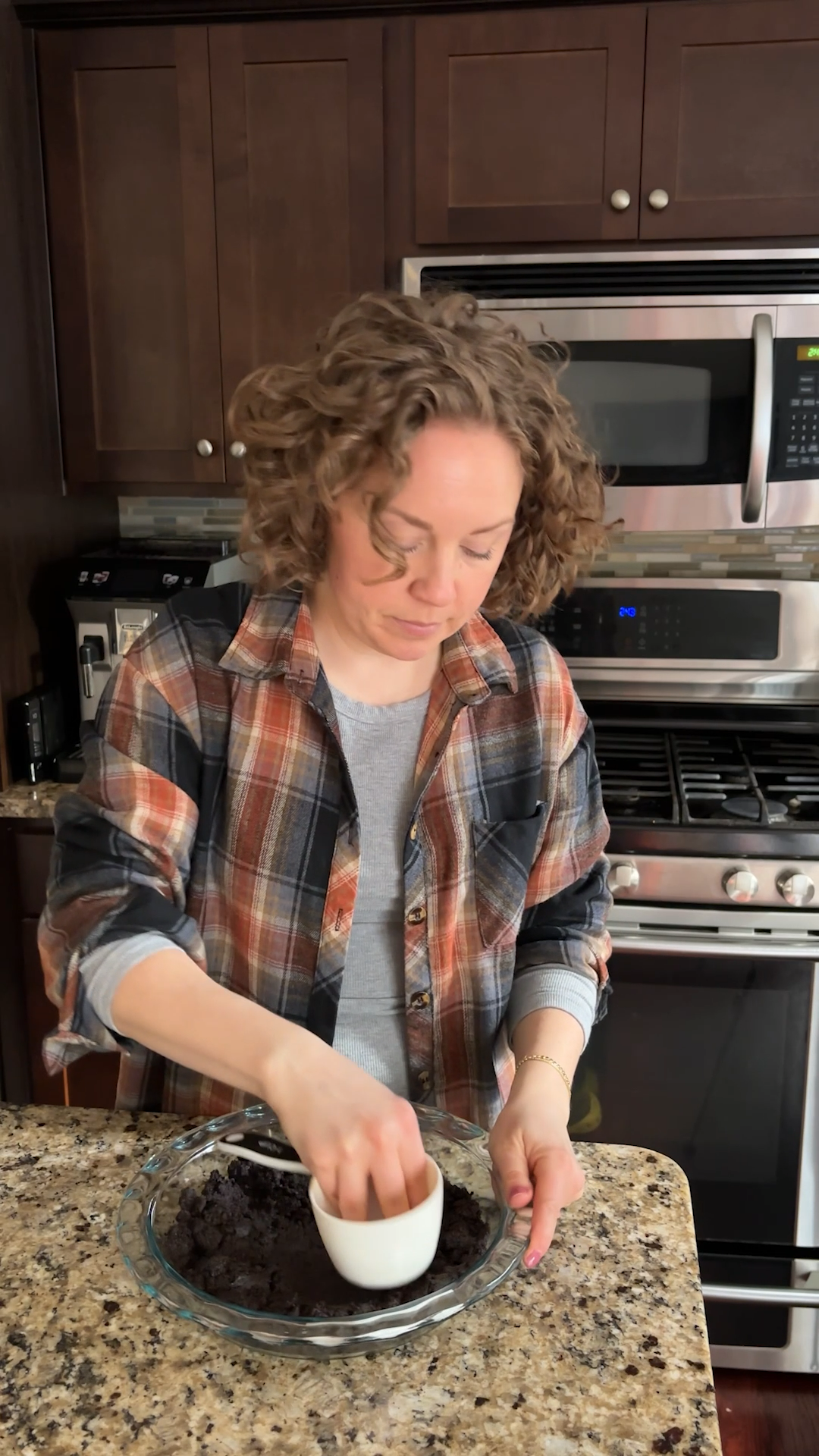 A woman with curly hair in a plaid shirt mixes ingredients for a no bake peanut butter pie in a bowl on a kitchen counter, with an oven and microwave in the background.
