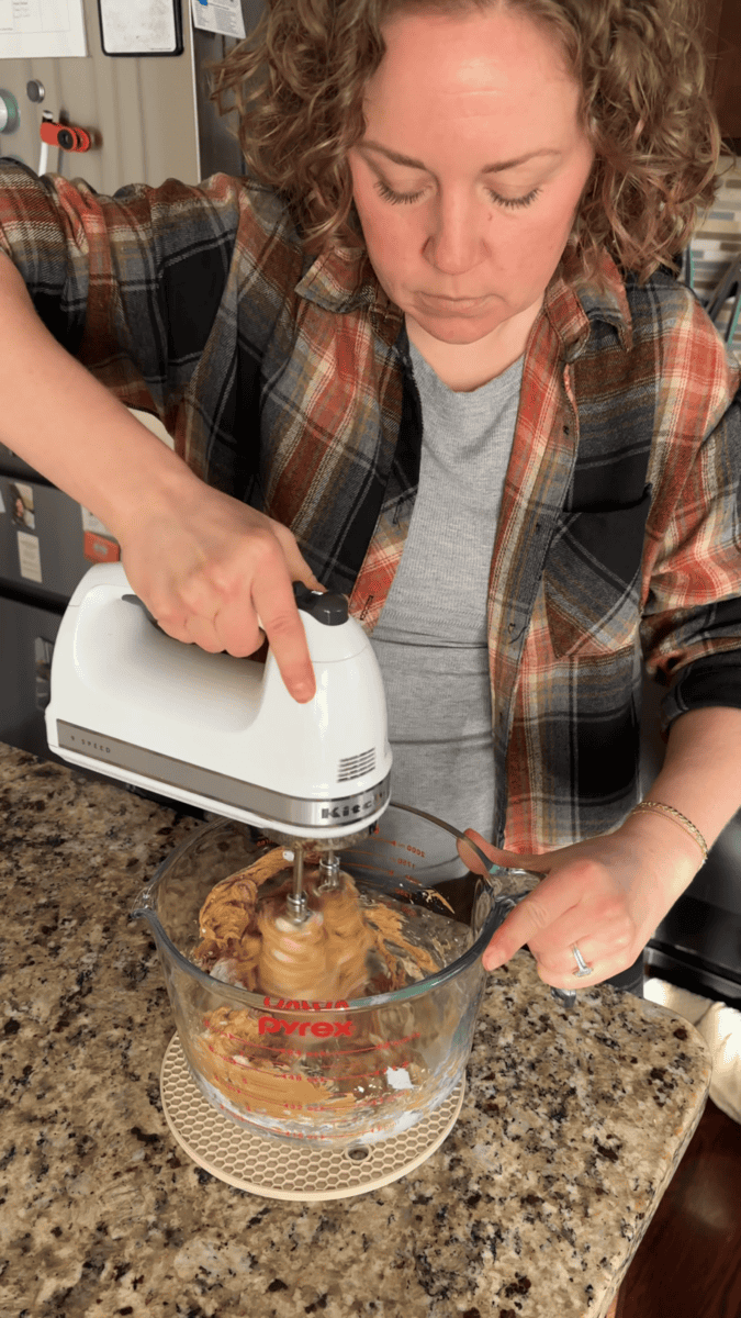 Person preparing a no bake peanut butter pie, using a white electric hand mixer to blend ingredients in a glass measuring bowl on a granite countertop, wearing a plaid shirt and gray tee.