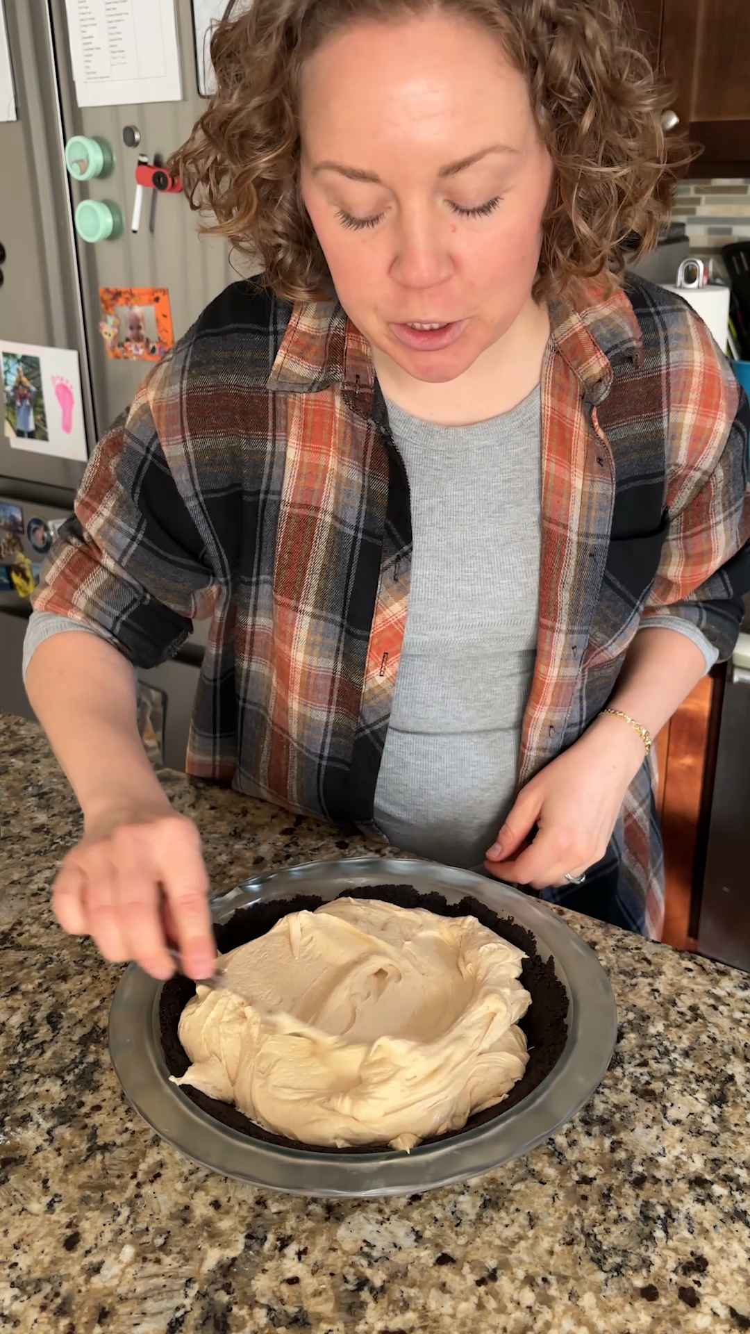 A person spreads a creamy filling for a no bake peanut butter pie into a pie crust on a granite countertop in a kitchen, wearing a plaid shirt and gray top.