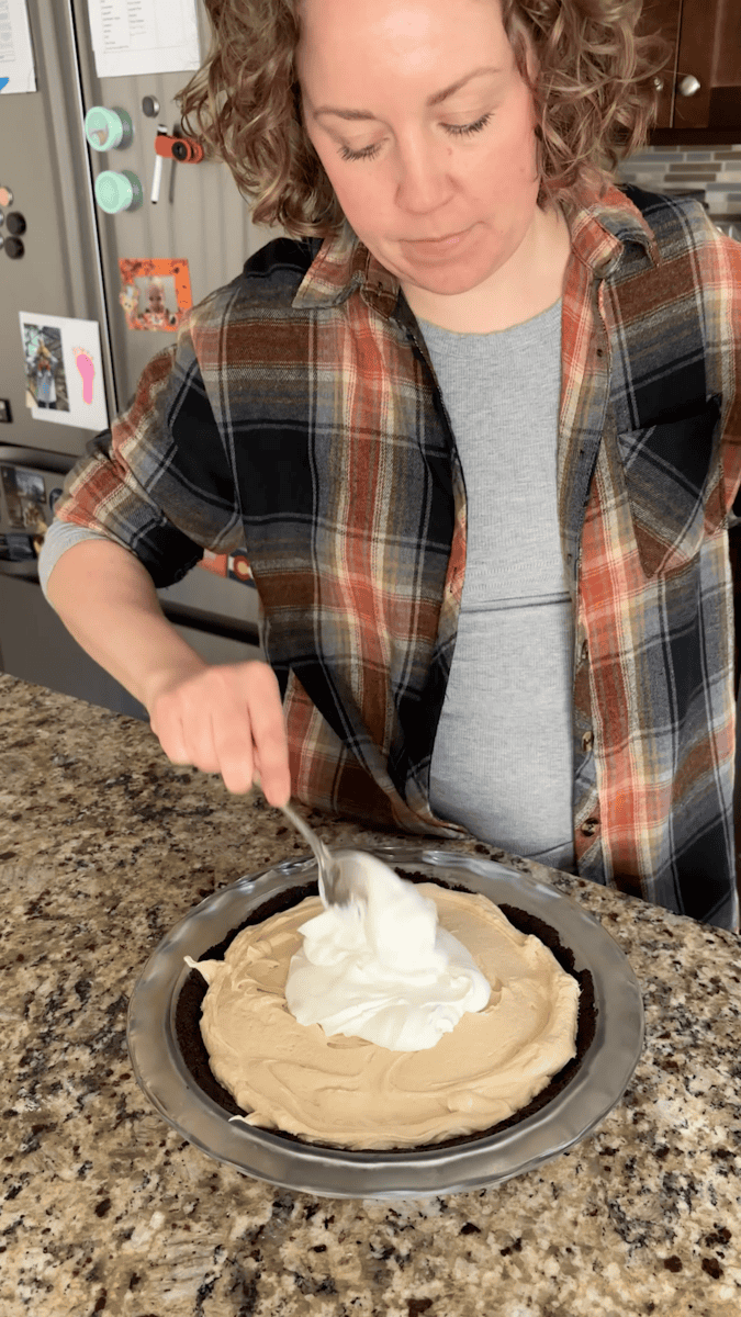 A person in a plaid shirt spreads whipped cream onto a no bake peanut butter pie with a spoon in a kitchen. The pie sits on a granite countertop near a refrigerator with colorful magnets and drawings.