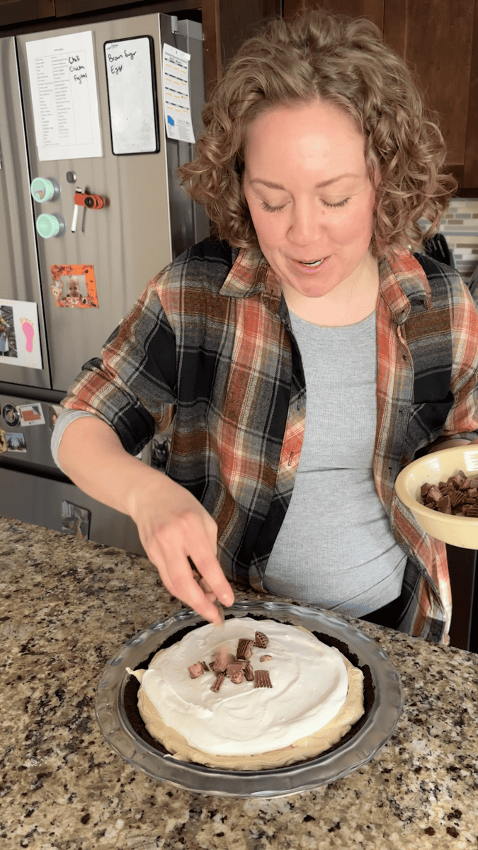 A person with curly hair tops a no bake peanut butter pie with chocolate pieces in a kitchen, holding a bowl and smiling while standing at a granite counter.
