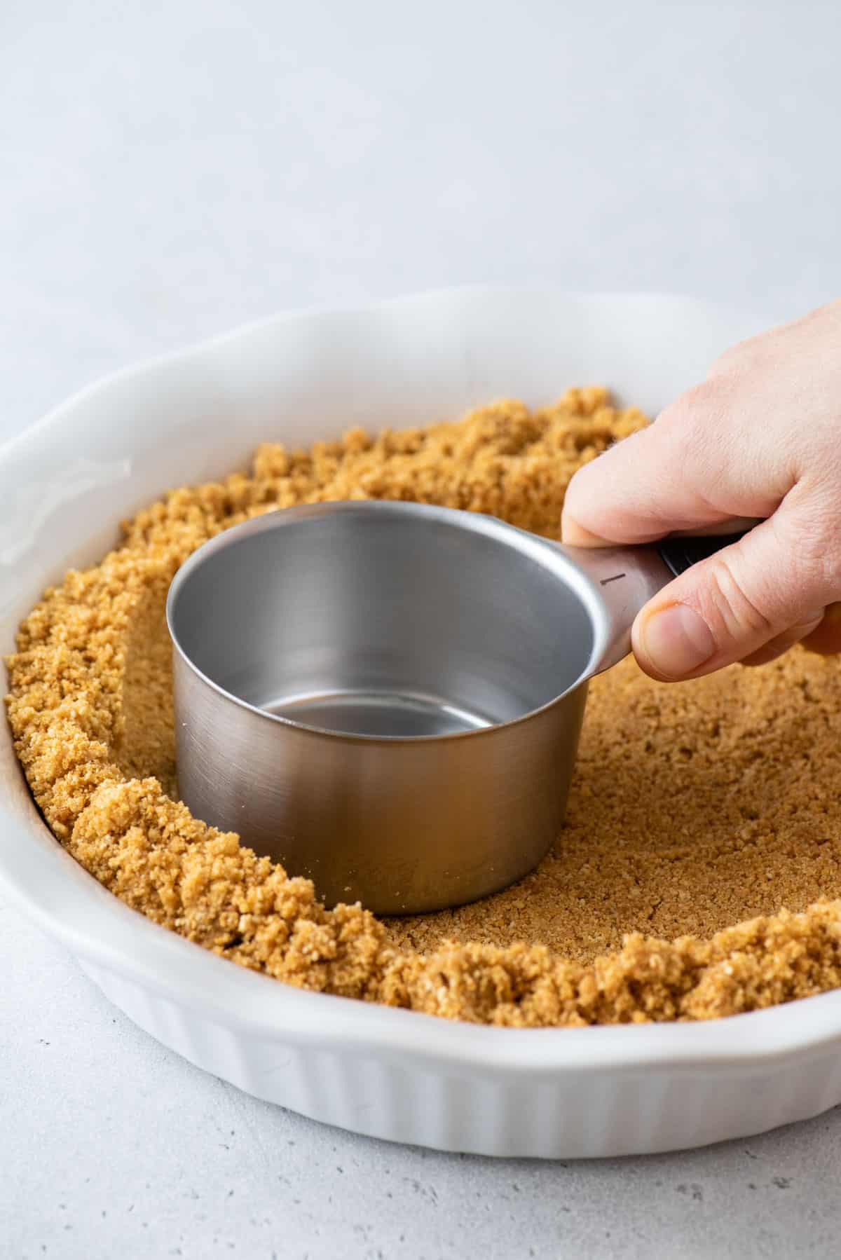 packing graham cracker pie crust into pie plate with metal measuring cup