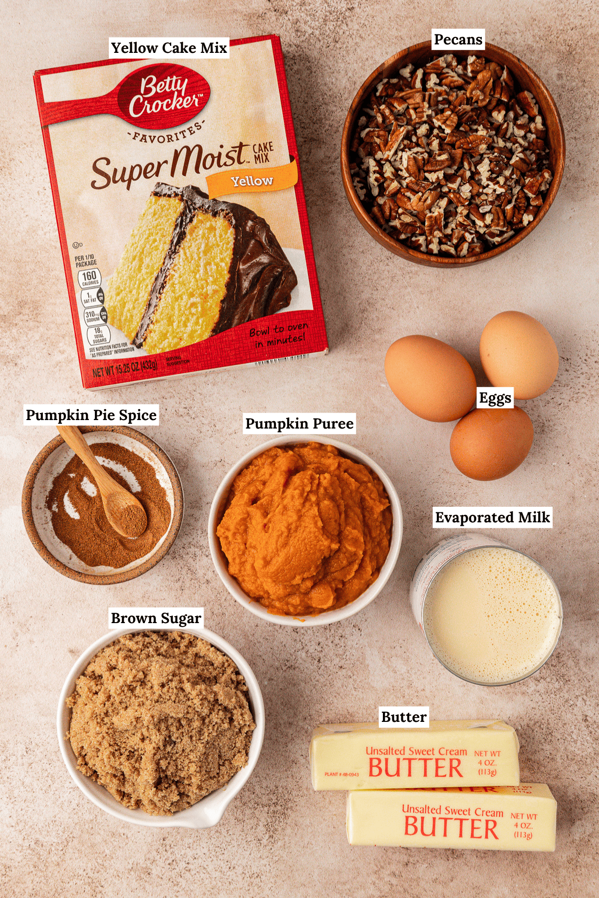 Ingredients for a pumpkin dump cake recipe displayed on a white surface: yellow cake mix, chopped pecans, eggs, evaporated milk, unsalted butter sticks, brown sugar, pumpkin puree, and pumpkin pie spice—each clearly labeled.