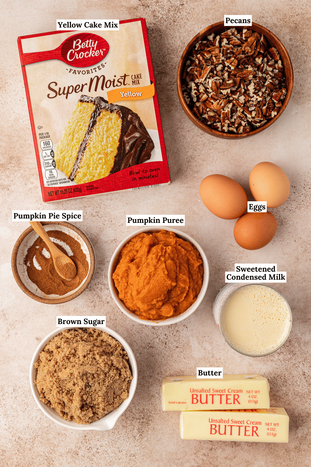 Labeled ingredients for a pumpkin dump cake recipe are arranged on a counter: yellow cake mix, chopped pecans, three eggs, pumpkin pie spice, pumpkin puree, sweetened condensed milk, brown sugar, and two sticks of unsalted butter.