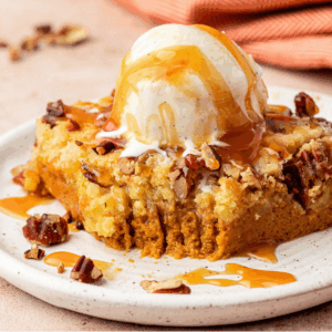A square slice of pumpkin dump cake topped with vanilla ice cream, chopped pecans, and caramel sauce sits on a white plate with a bite taken out. A striped orange napkin is in the background.