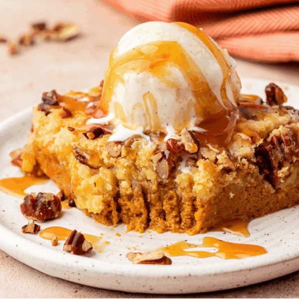 A square slice of pumpkin dump cake topped with vanilla ice cream, chopped pecans, and caramel sauce sits on a white plate with a bite taken out. A striped orange napkin is in the background.