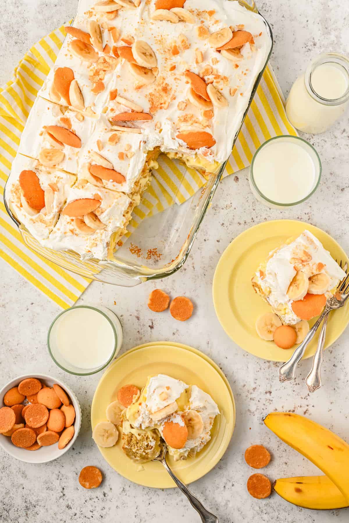 partially eaten banana poke cake in a baking dish sitting on a white and yellow striped towel on a counter with glasses of milk, slices of cake on plates, and vanilla wafers arranged around it