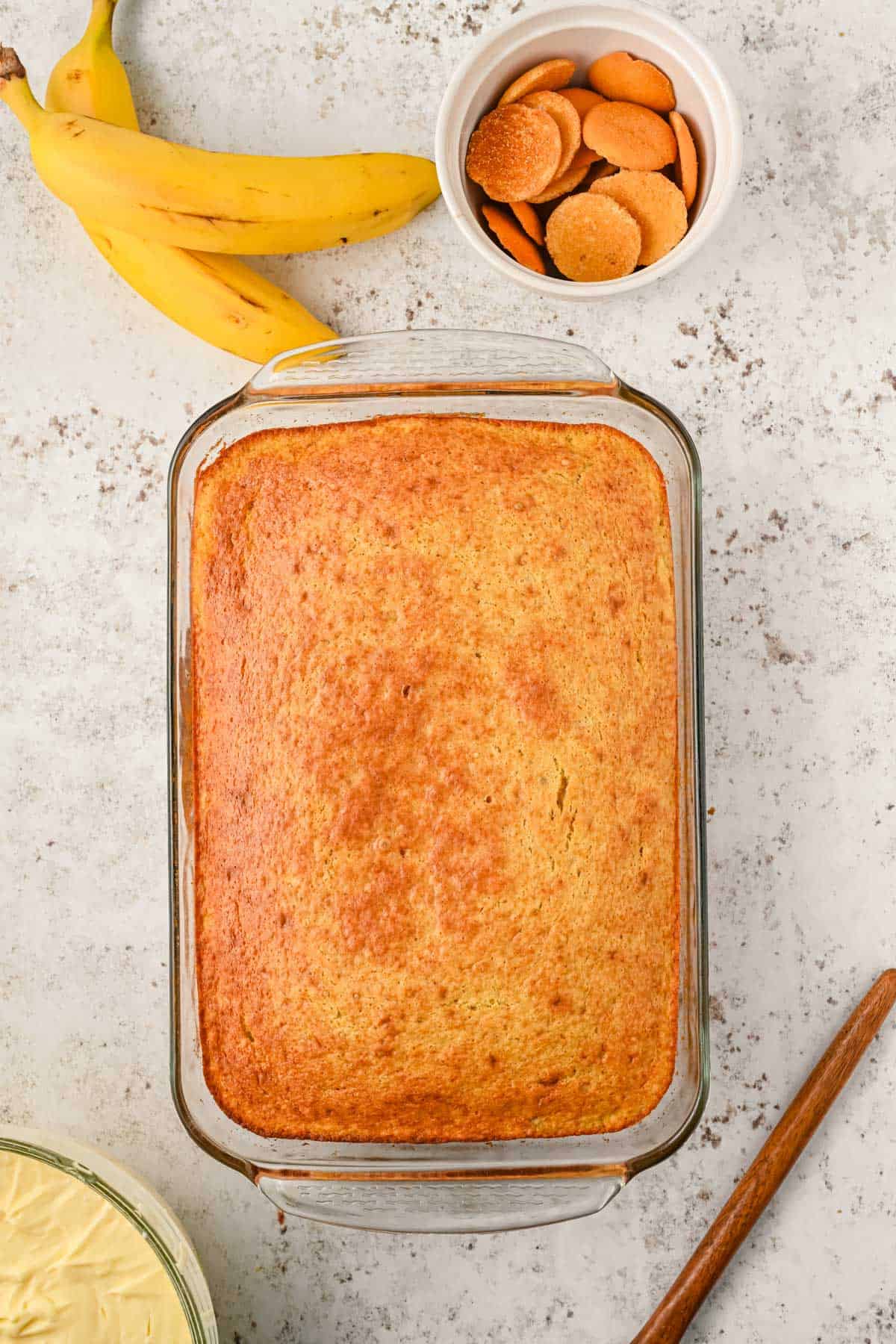 banana cake in a clear baking dish on a counter with a wooden spoon and bowl of banana pudding beside it