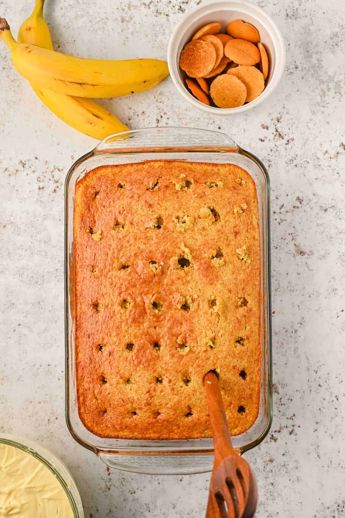 banana cake in a clear baking dish on a counter with a bowl of banana pudding beside it and a wooden spoon being used to poke holes in it