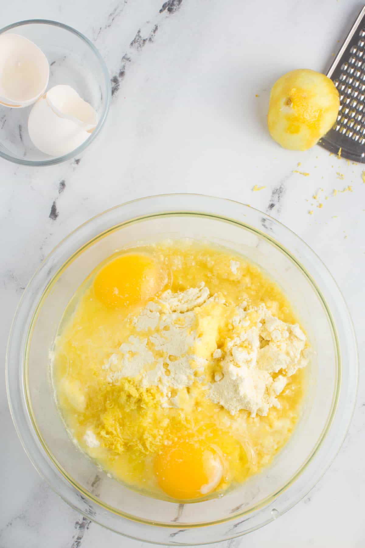 ingredients for lemon cookies in a bowl partially mixed, sitting on a counter top with lemons, a grater, and empty egg shells around it.