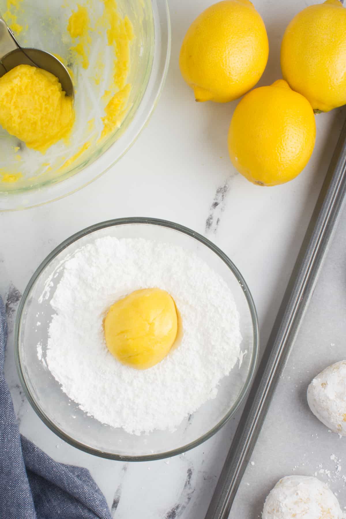 an arrangement of lemons, lemon cookie dough in a bowl, and powdered sugar in a bowl with a lemon, sitting on a baking sheet lined with parchment paper