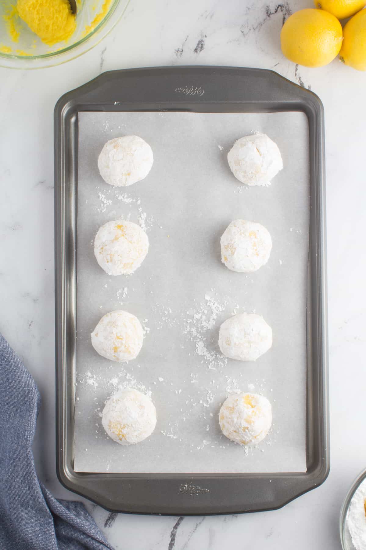 lemon cookie dough balls coated in powdered sugar sitting on a baking sheet lined with parchment paper