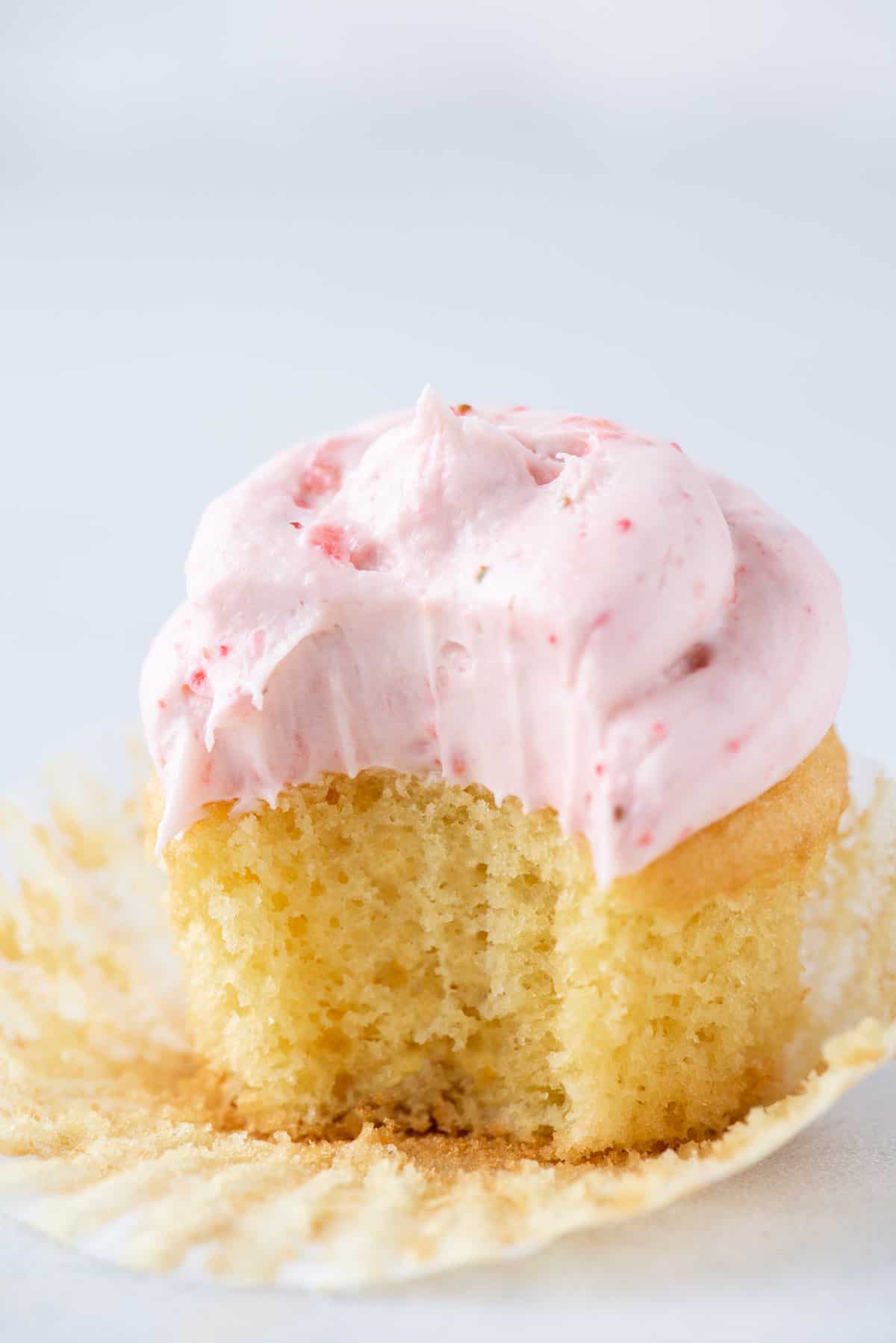 partially eaten vanilla cupcake with strawberry frosting sitting on an open paper muffin cup