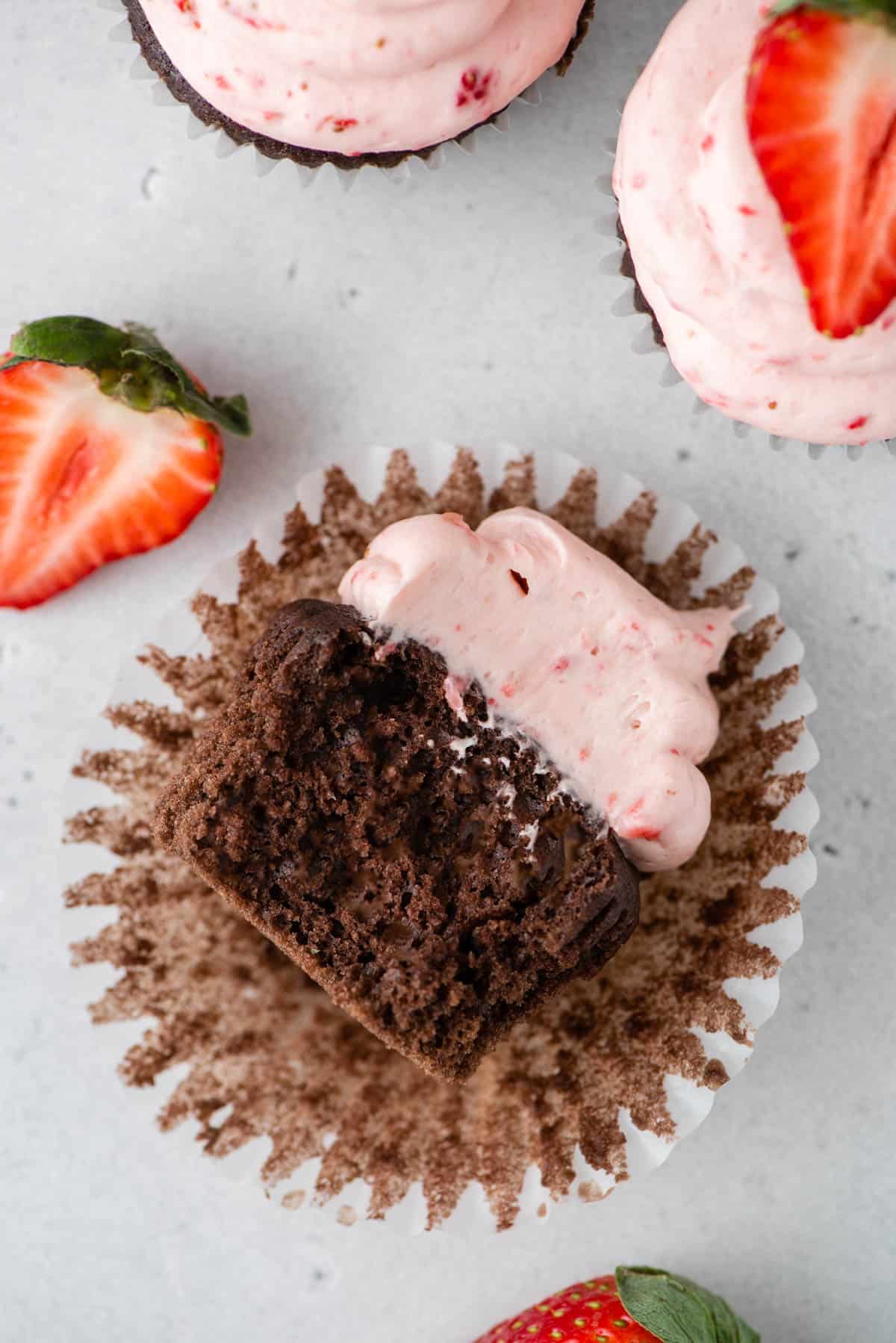 partially eaten chocolate cupcake with laying on it's side on a paper muffin cup with fresh strawberries and more cupcakes arranged around it
