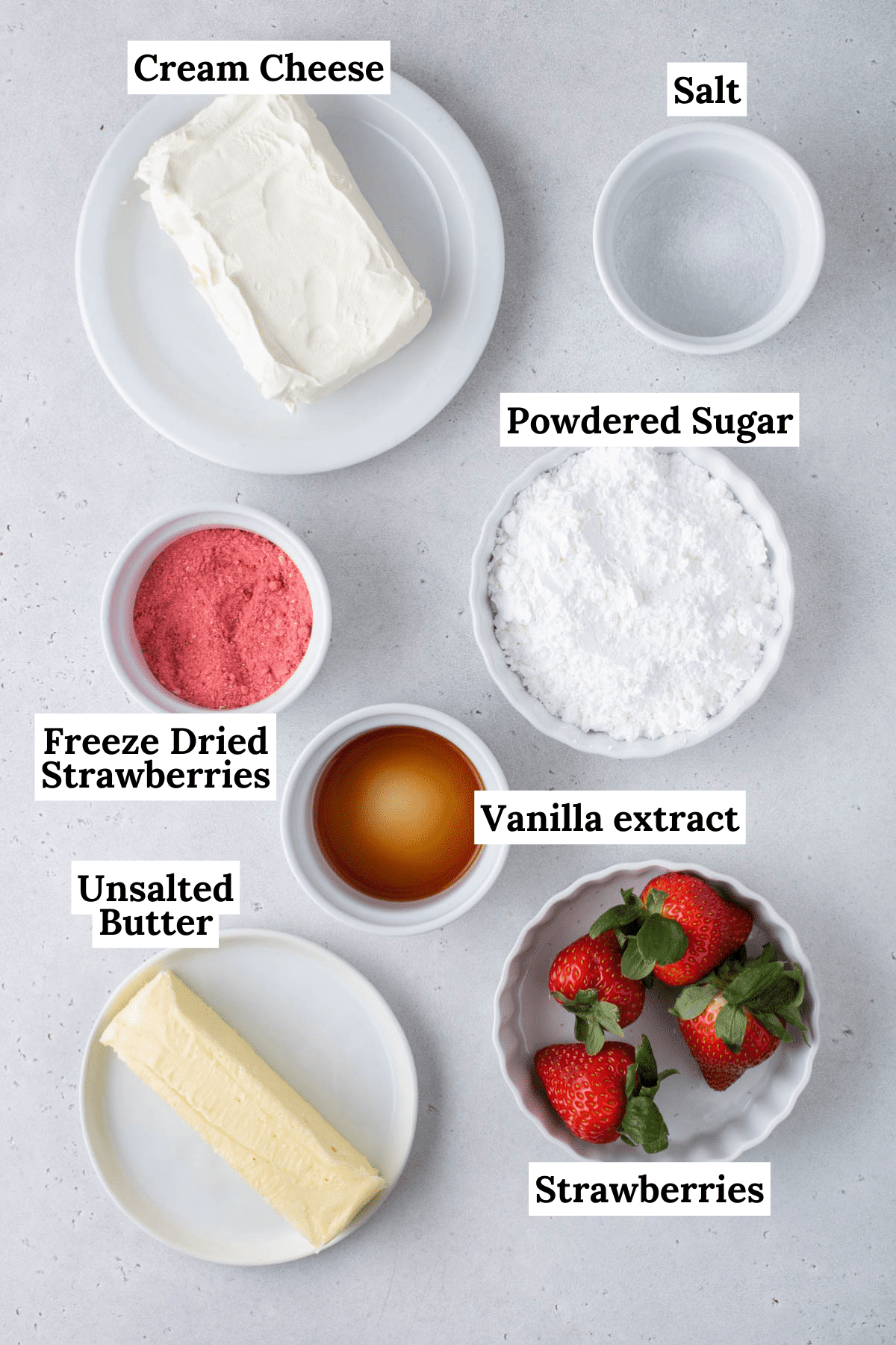 ingredients for strawberry cream cheese frosting in separate white bowls including a stick of butter, fresh strawberries, vanilla extract, powdered sugar, ground freeze fried strawberries, cream cheese and salt