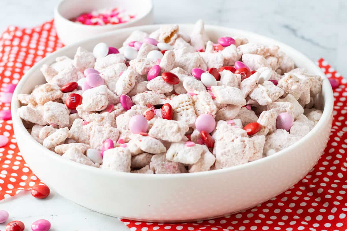 Valentine's Day Puppy Chow in a white bowl sitting on top of a white counter and red napkins with white polka dots on them.