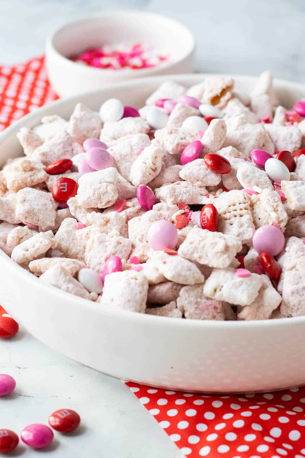 Valentine's Day Puppy Chow treat in a white bowl sitting on top of red and white napkin with pink & red m&ms sprinkled around it.