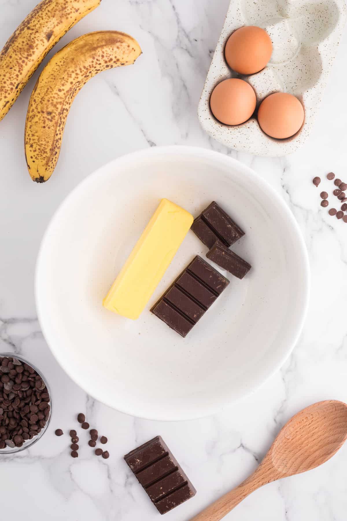butter and semi sweet chocolate in a bowl surrounded by bananas, eggs, a wooden spoon and chocolate chips