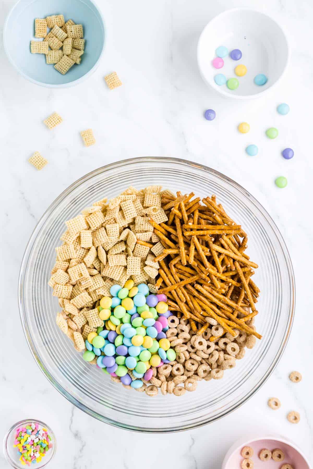 dry ingredients for bunny bait in a clear glass bowl surrounding by a bowl of sprinkles, a bowl with cheerios in it, a bowl of rice chex and a bowl of m&ms