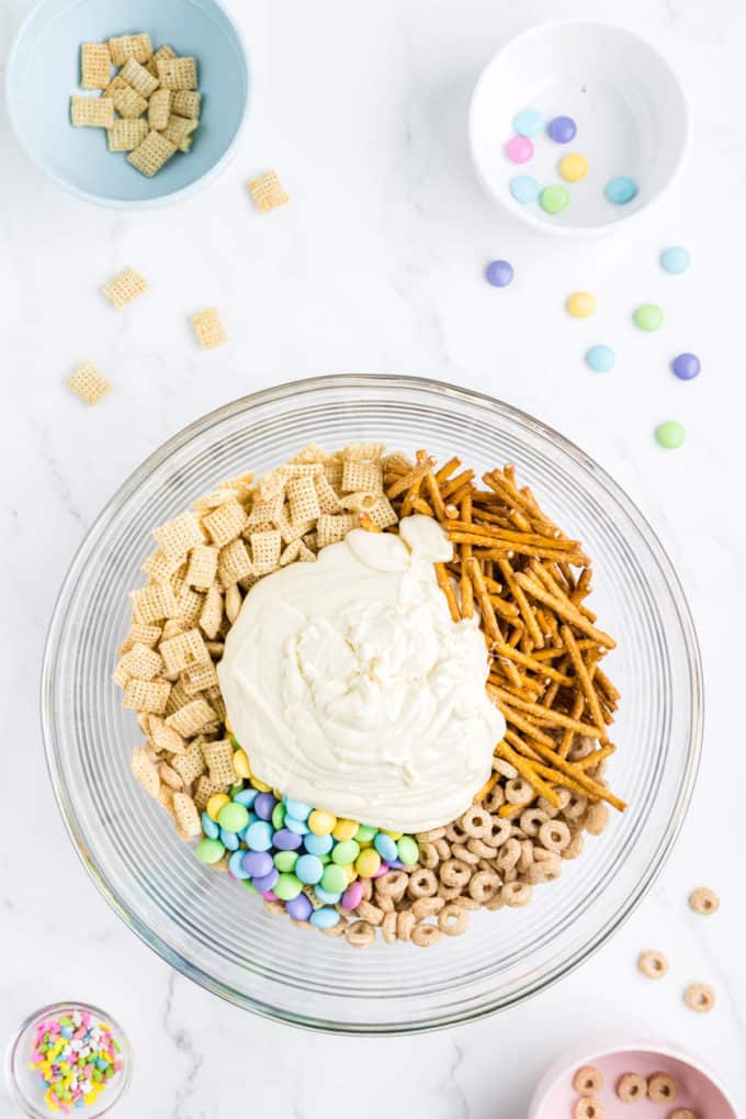 ingredients for bunny bait in a clear glass bowl surrounding by a bowl of sprinkles, a bowl with cheerios in it, a bowl of rice chex and a bowl of m&ms